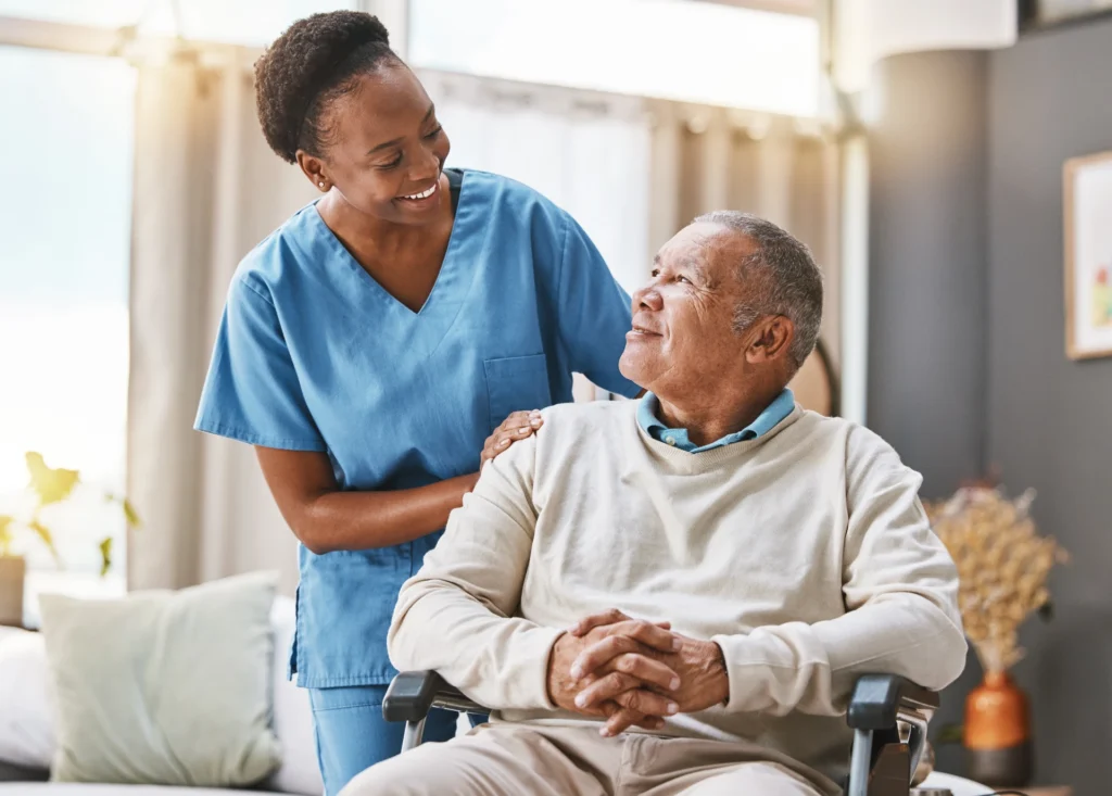 Female nurse smiles down at elder patient who is sitting in his wheelchair.