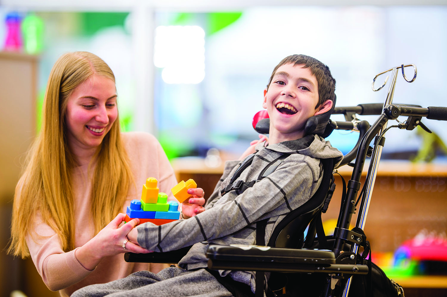 Very happy, smiling male pediatric patient in a wheelchair with a caregiver holding blocks in front of his wheelchair
