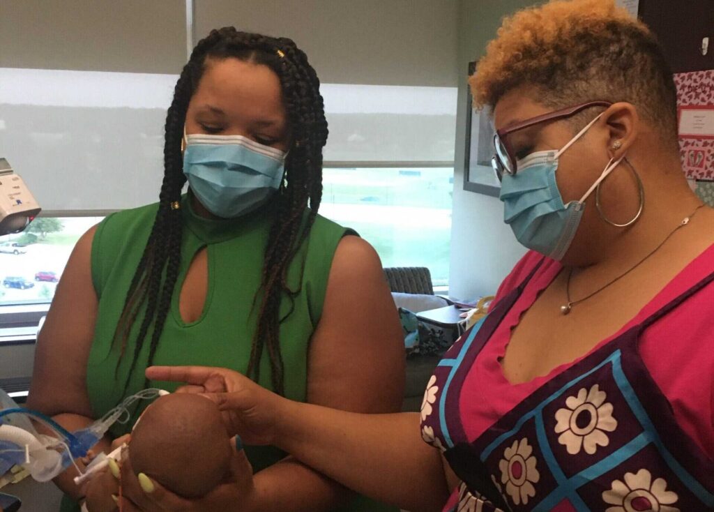 Aunt Erin holding baby Devyn in the NICU while Elana, her mother, touches her face. Both sisters are wearing face masks.