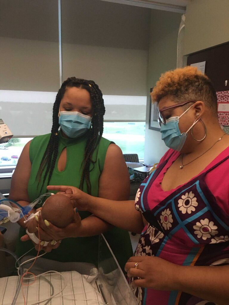 Aunt Erin holding baby Devyn in the NICU while Elana, her mother, touches her face. Both sisters are wearing face masks.