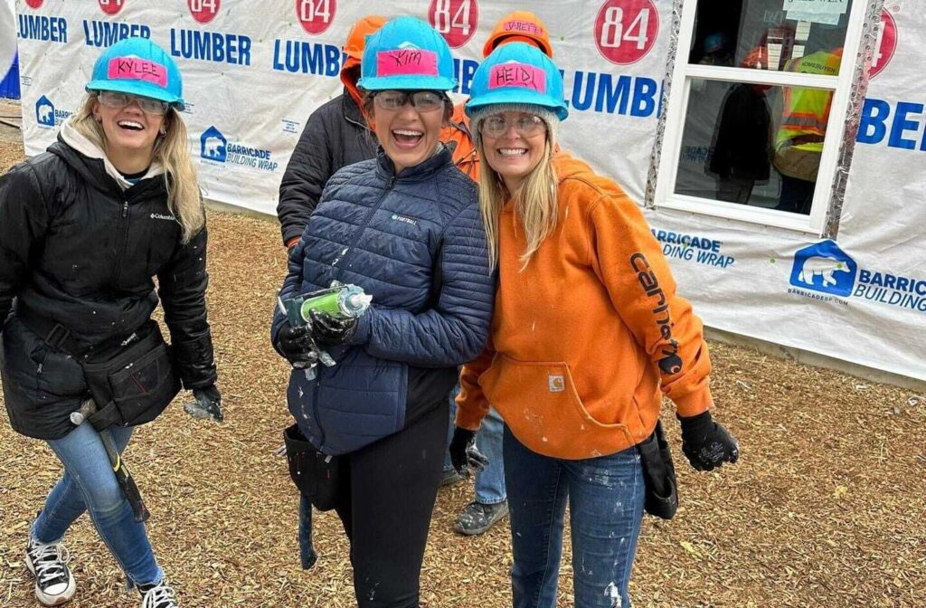 Kylee, Kim, and Heidi smile and laugh while holding caulk in front of the Habitat for Humanity home being built. They're dressed to keep warm and wearing hard hats.