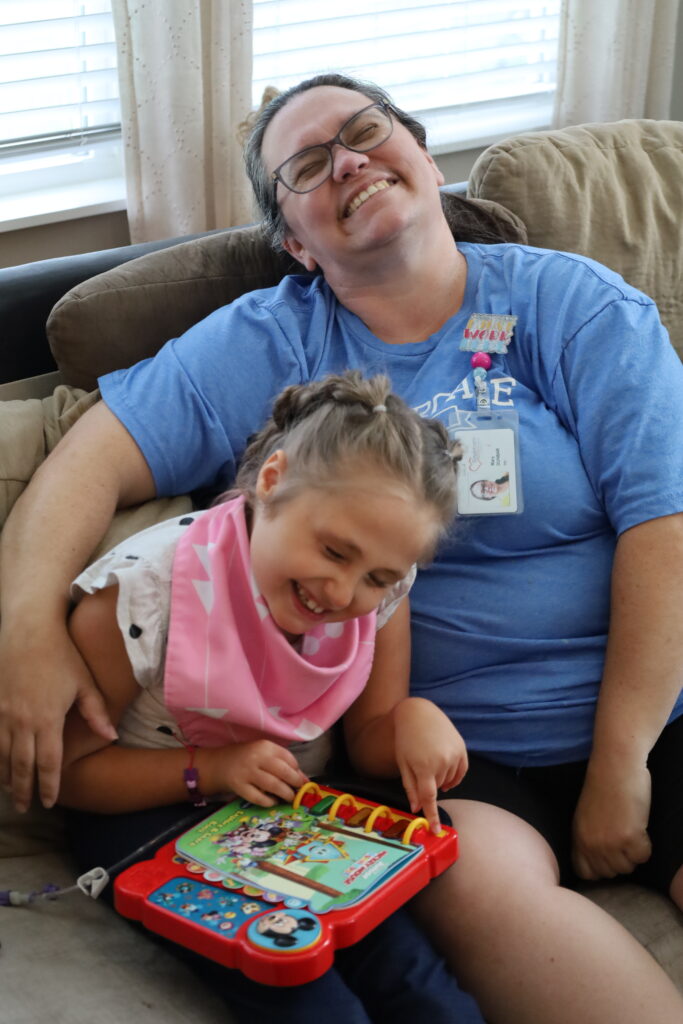 Ruby and caregiver Mary sitting on a couch, both smiling. Ruby is playing with a colorful piano toy, she loves music!