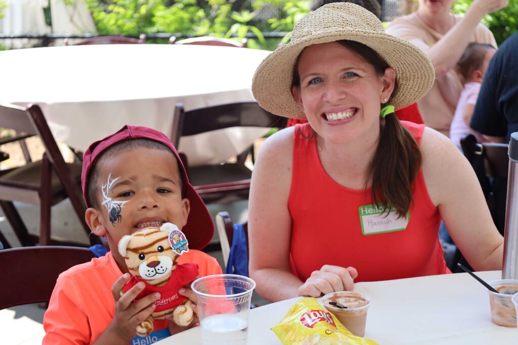 Hannah is wearing a red shirt with a straw hat and sits with a child who has a spider facepaint and is holding a tiger who has a red Tendercare shirt on.