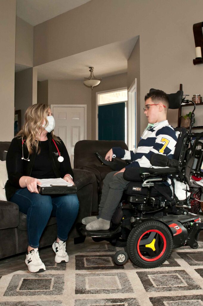 A healthcare worker in a mask and scrubs sits on a couch, talking to a person in a motorized wheelchair inside a home.