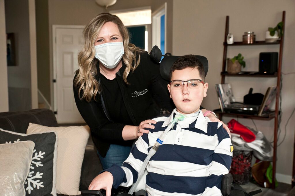 A nurse wearing a face mask stands beside a boy in a wheelchair. The boy wears glasses and a striped shirt, with a neck brace and tracheostomy tube visible. They are in a living room with a couch, decorative pillows, and a shelf with books and plants in the background.