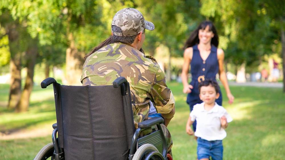 Army veteran is pictured facing away from the camera and toward his young family who is running toward him in his wheelchair.